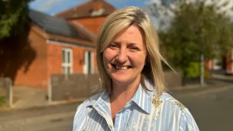 A woman with blonde hair, wearing a blue and white stripy shirt, smiling at the camera. She is standing across the road from a row of red brick homes. 