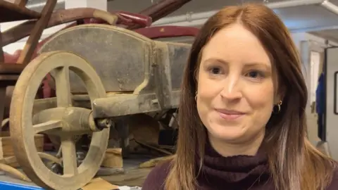 BBC Helen is wearing a dark maroon sweater and standing indoors. Behind her is an old wooden cart with large spoked wheels.