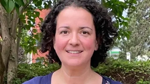 A woman with dark curly hair smiles at the camera, pictured from the shoulders up. She wears a purple top and has trees behind her.
