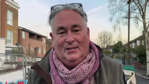 A man in a pink patterned scarf, green gilet  and glasses on his head looks at the camera. Behind him are houses and temporary works fencing.