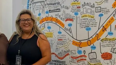 Safiya Saeed Myroslava Coates with long blonde hair, wearing a black vest stop and large black rimmed glasses, standing in front of her artwork. 