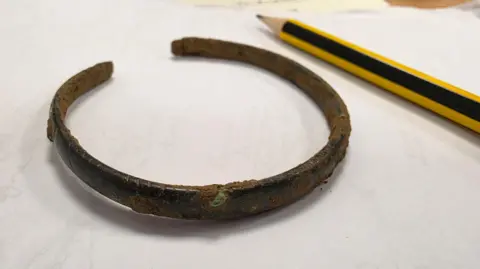 University of Leicester A copper-alloy late Roman pennanular bracelet, on a piece of paper, with a pencil to the right.