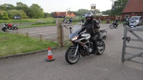 Steve Jones A slightly out of focus images of Steve Jones in black leathers on a black motor bike as he drives through a gate. Behind him is carpark with a few parked up motorbikes, green fields and a a couple of red brick buildings. 
