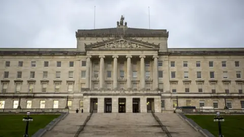 PA Media A wide shot of the exterior of Stormont/Parliament Buildings. It features a large, white-stone building with six large columns in the front with a large, wide staircase leading up to the doors.