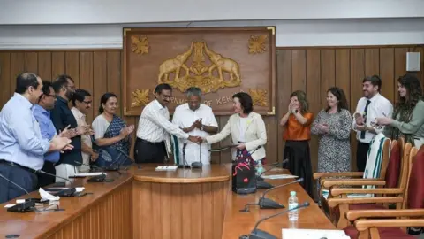 Welsh government Eluned Morgan, when she was the Health minister,  and a Welsh government delegation meeting meeting there counterparts in Kerala India they are at the had of a push wooden board room with a wood engraved placard on the wall above them. 