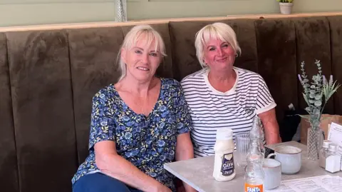 Two women, both with blonde hair, sitting together and smiling at a coffee shop. One wears a blue flowery top while the other is in a white and navy-striped t-shirt.