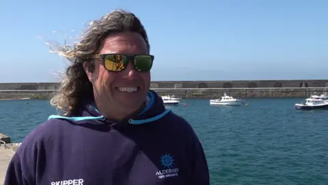 Dan Slimm is smiling at the camera as he stands on the quayside on a bright sunny day. He has shoulder-length curly hair and is wearing sunglasses. His hoodie is branded and reads SKIPPER ALDERNEY FERRY SERVICES. Behind him are four small boats moored inside the harbour.