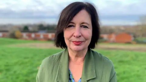 A woman with dark hair and a green jacket standing on a grassy area with blurred houses in the background