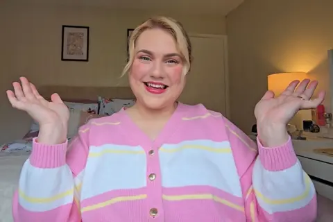Isobel Guffick Isobel Guffick, a blonde woman in a pink, white and yellow cardigan, sits in a bedroom and smiles at the camera with her hands in the air