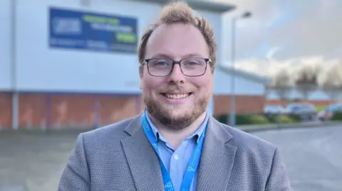A man with fair hair and beard, wearing a grey blazer and blue lanyard, standing in front of a white cladded building with a brick base