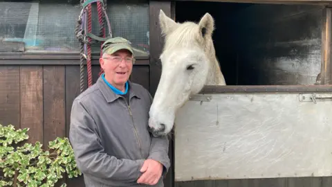 LDRS Smiling Alan French, wearing a cap and brown jacket, stands next to a white horse in its stable