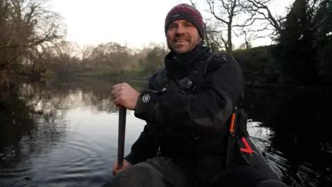 Dan Smith sitting in his canoe on the River Tees. He is wearing a dark-coloured bobble hat and wearing a black waterproof jacket, a black lifejacket and black trousers and is holding a paddle in two hands to the side of him