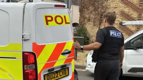 A police officer wearing black uniform and Police written on the back of his shirt.
He is standing next to a police van and the back door slightly open - he appears to be closing the door of the van. There is a white car on the right hand side parked on the road.