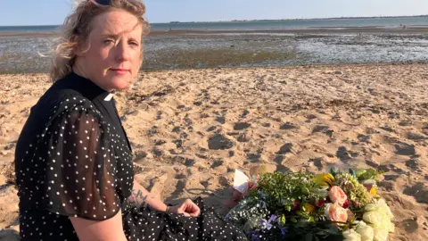 Henry Godfrey-Evans/BBC The Reverend Jo Parrott wears a black and white spotted dress with a white clerical collar. She is sitting on a sandy beach with flowers on the sand. The sea is in the distance.