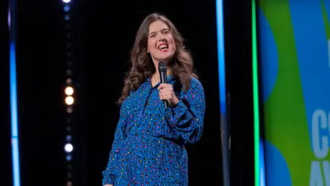 BBC/Phil McIntyre Television/Michael Prince Comedian Rosie Jones wearing a blue leopard print jumpsuit. She is smiling with her mouth open and holding a black microphone. She appears to be on stage with a black, blue and green backing behind her.