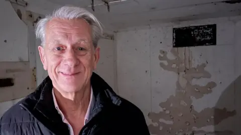 Andrew Turner/BBC Trevor Blythe, in a black jacket and pink shirt, standing in the bunker, and the ventilation shaft to the left of the image.