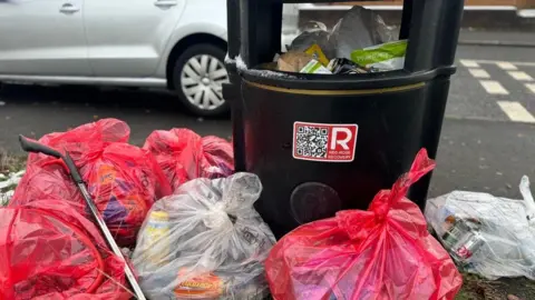 A rubbish bin ins full and surrounded by tied bin bags full of rubbish.