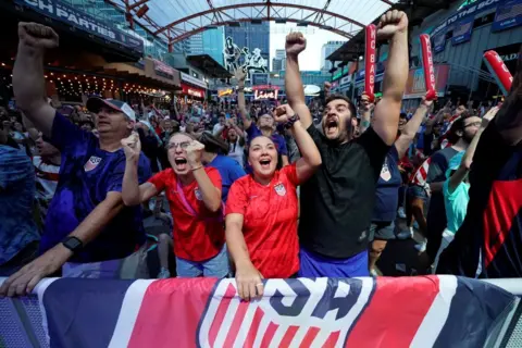 Fans dressed in USA shirts and with USA flags cheer with arms aloft
