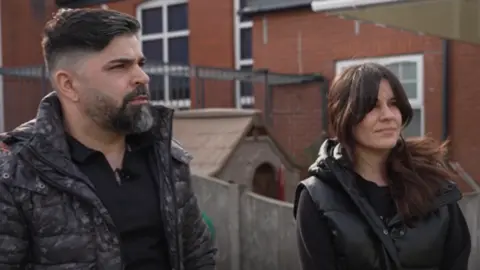 Alice's dad Sergio, wearing a black top and dark grey jacket, stands next to his wife Alex, who is also wearing black, in their daughter's old school playground in Southport.