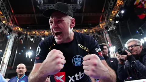 BRADLEY COLLYER / PA WIRE Tyson Fury celebrates, he wears a black t-shirt and cap as he clenches his fists in a sports arena, people take pictures of him in the background.