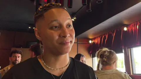 Kay Greyson looking to the right of the camera with pursed lips. They have closely dark, shaved hair on the sides with braids on the top. They are wearing two gold necklaces and a black v-neck t-shirt. There are people standing behind them and windows to the right.