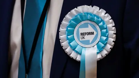A close up picture of a Reform UK blue and white rosette attached to a suit.