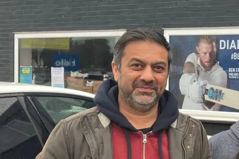 ALEXANDER STEVANOVIC/BBC Ray is standing outside his cricket training business. There is a car to the left of the picture and a poster in the background.