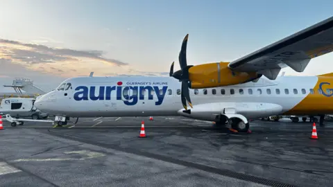 An Aurigny plane seen on the runway. It is white with yellow as its secondary colour and large black propellers. 