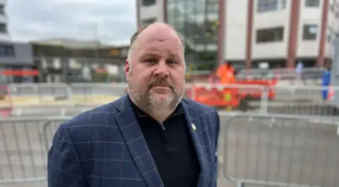 A man in a suit jacket and navy jumper looks at the camera. He is standing outside and there are barriers behind him.