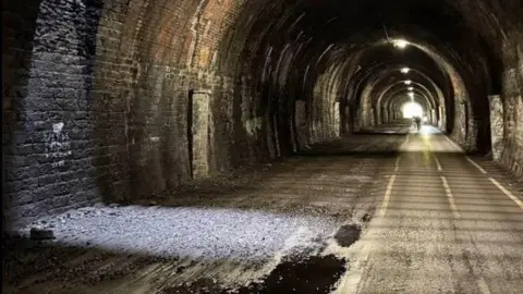 Google A tunnel section of the Bristol to Bath Cycle Path. The inside is lit up by a few lights on the ceiling and a couple of cyclists can be seen approaching the camera.