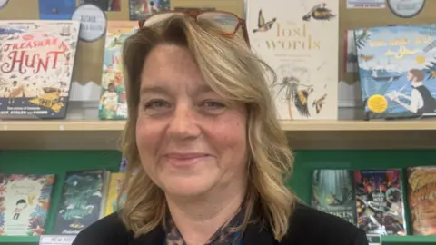 Annabel Amos/BBC Julie Broadbent is in the centre of the image looking at the camera. She has shoulder length dark blonde hair and is wearing a black jacket. She is sat in front of children's books on a bookshelf. 