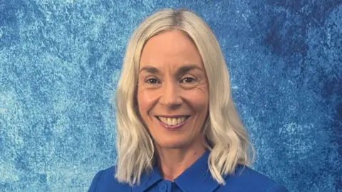 Malone Integrated College Aine Leslie. She has shoulder-length blonde hair and brown eyes, wearing a blue buttoned-up jacket. Behind her is a blue background. She is smiling at the camera.