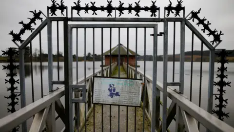 A sign hangs on a metal gate at Severn Trent Water's Cropston Reservoir. A bridge can be seen leading towards a brick building. 