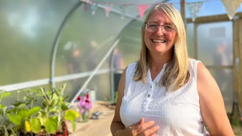 Sheena Brockie smiles while standing inside a polytunnel which is being used by not-for-profit organisation Grow to run classes all year round. She has shoulder length blonde hair and is wearing a white sleeveless top. She is stood next to a wooden table with several small plants on it. People can also be seen standing outside the polytunnel through its opaque panels.