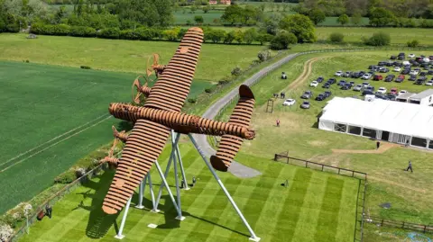An aerial view of a large, brown‑coloured metal sculpture shaped like a plane mounted on angled support legs in the middle of a neatly mown field. The surrounding area includes open green fields, a curving path and a rural landscape stretching into the distance under a cloudy sky. To the right, a grassy car park filled with vehicles and a long white marquee suggests an outdoor event, with several people walking around the site.
