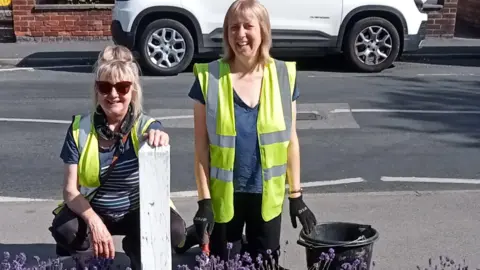 Two women smiling on a sunny day in a residential street with lavender growing in front of them. They are both wearing yellow hi vis vests. The woman on the left is squatting with her hand on a short white wooden post and the woman on the right is kneeling down, wearing black gloves with one hand resting on a black bucket.