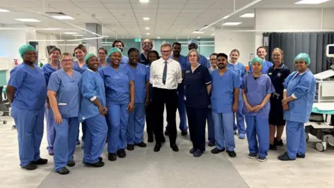 A group picture on the ward. Hospital staff are wearing blue scrubs, Mr Wood is wearing a white shirt, black trousers and a tie.