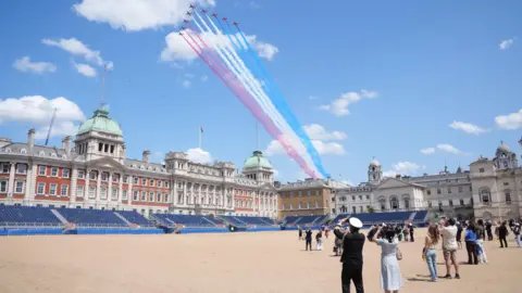 PA Media People watch on from the parade ground as the Red Arrows streak past overhead trailing red, white and blue smoke