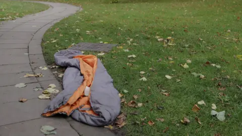 An empty grey and orange sleeping bag lies on a grassed area covered with autumn leaves. A footpath runs alongside the grass.