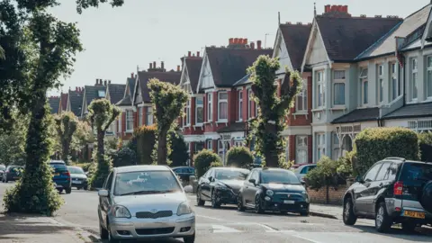 Getty Images A quiet suburban street with cars parked outside houses and trees lining both sides of the street. 