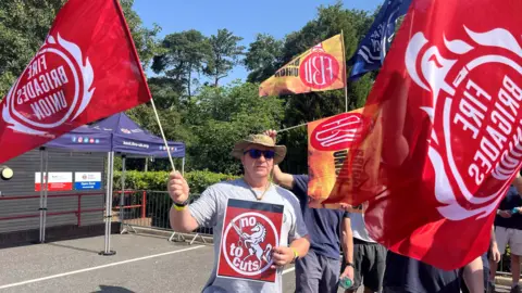 BBC A man in a wide-brimmed hat waves a red flag and holds a placard protesting cuts