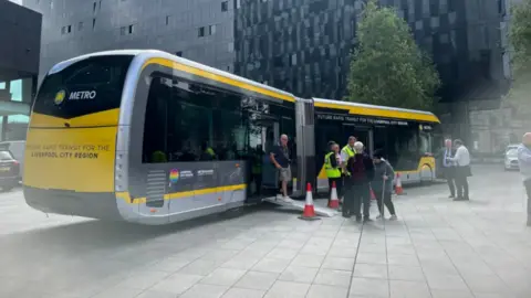 Image shows a low-sitting, articulated bus liveried up in yellow with the word 'Metro' on it. A number of people are standing around looking at it. 