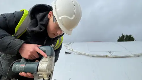 Family handout Jack busy at work, wearing a hard hat, hi-vis vest over a black puffer jacket.  He is using an electric jigsaw appliance 