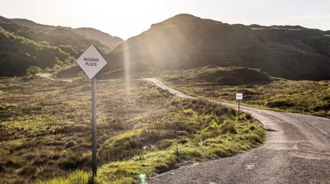A rural landscape with a tarmac road running through hills. There are signs indicating passing places for vehicles. The sun is shining.