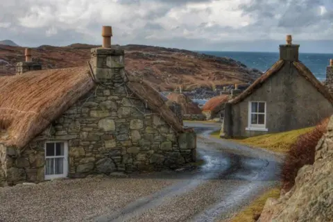 John Dyer Some stone built houses sut into the ground. They have thatched roofs. A road twists through them and is curiously at a height that is half way up the houses.