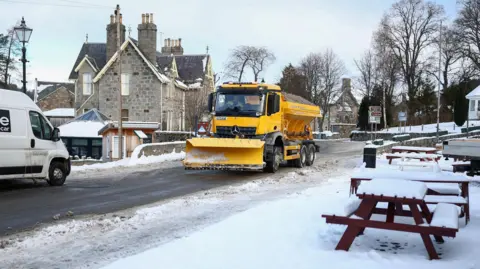 Getty Images A yellow gritting lorry fitted with a snow plough travels down a snowy street in the picturesque village of Braemar.