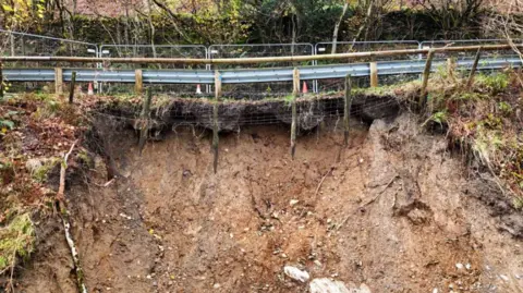 Westmorland and Furness Council Close up shot of the side of the road with a grey fence in the foreground and trees in the background. There are orange cones by the fence. Underneath the fence is exposed soil which shows signs of a significant landslip. 