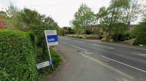 A street view of a suburban road with greenery on both sides. In the foreground, there is an estate agent's sign reading ‘Manning Stainton – Sold’ and a smaller street sign which says ‘Green Lane’.