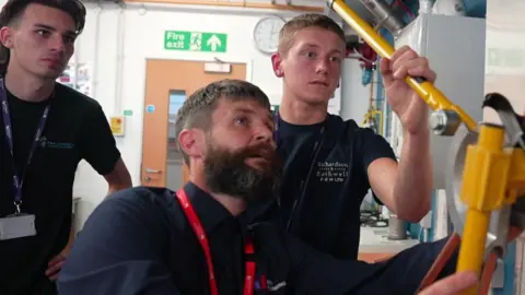 Richard Knights/BBC A man with short hair and a black beard is with two young male students as one of them tries to bend a pipe using a piece of equipment.