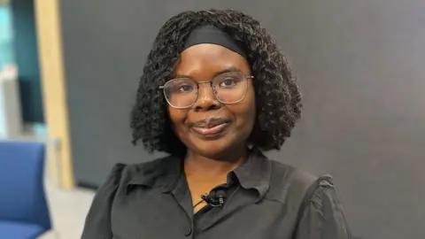 Emma Baugh/BBC Young woman with a head band wearing a black top in front of a black background. She is wearing glasses and has a smile on her face
